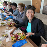 A group of women seated along a long table learning to create a charcuterie board with grapes, crackers, cheese, and fruit in a bright indoor event space.